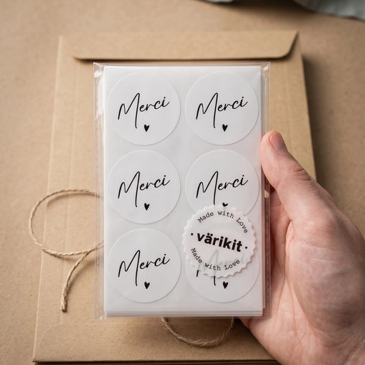 Hand holding a clear cellophane sleeve containing a sheet of Handwritten-style typography stacked with a single small solid heart Varikit stickers reading 'Merci'.