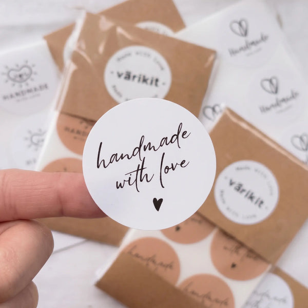 Hand holding a Handwritten-style typography stacked with a single small solid heart Varikit sticker reading 'handmade with love', with wrapped sticker sheets and branding materials in the background.