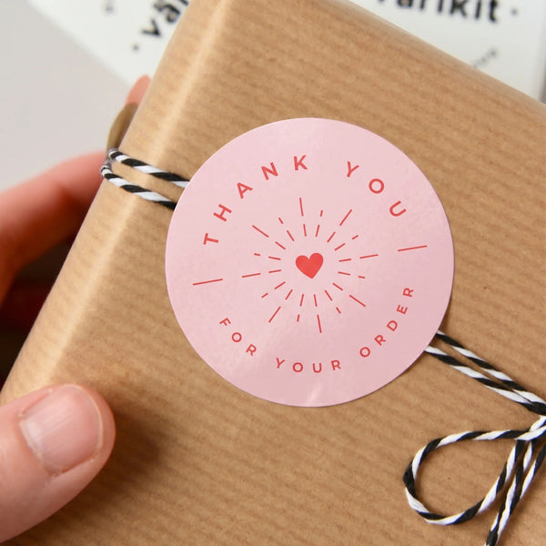 Hand holding a kraft paper package tied with black and white twine, featuring a Thank You Radiant Starburst Varikit sticker reading 'Thank You'.