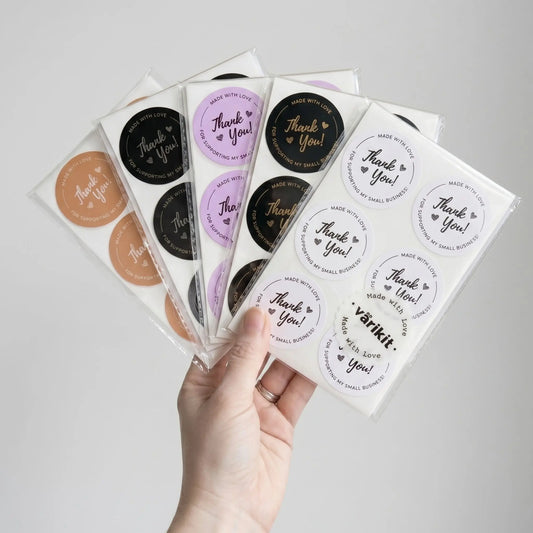 Hand holding several sheets of round stickers with 'Thank You' text on a light gray background