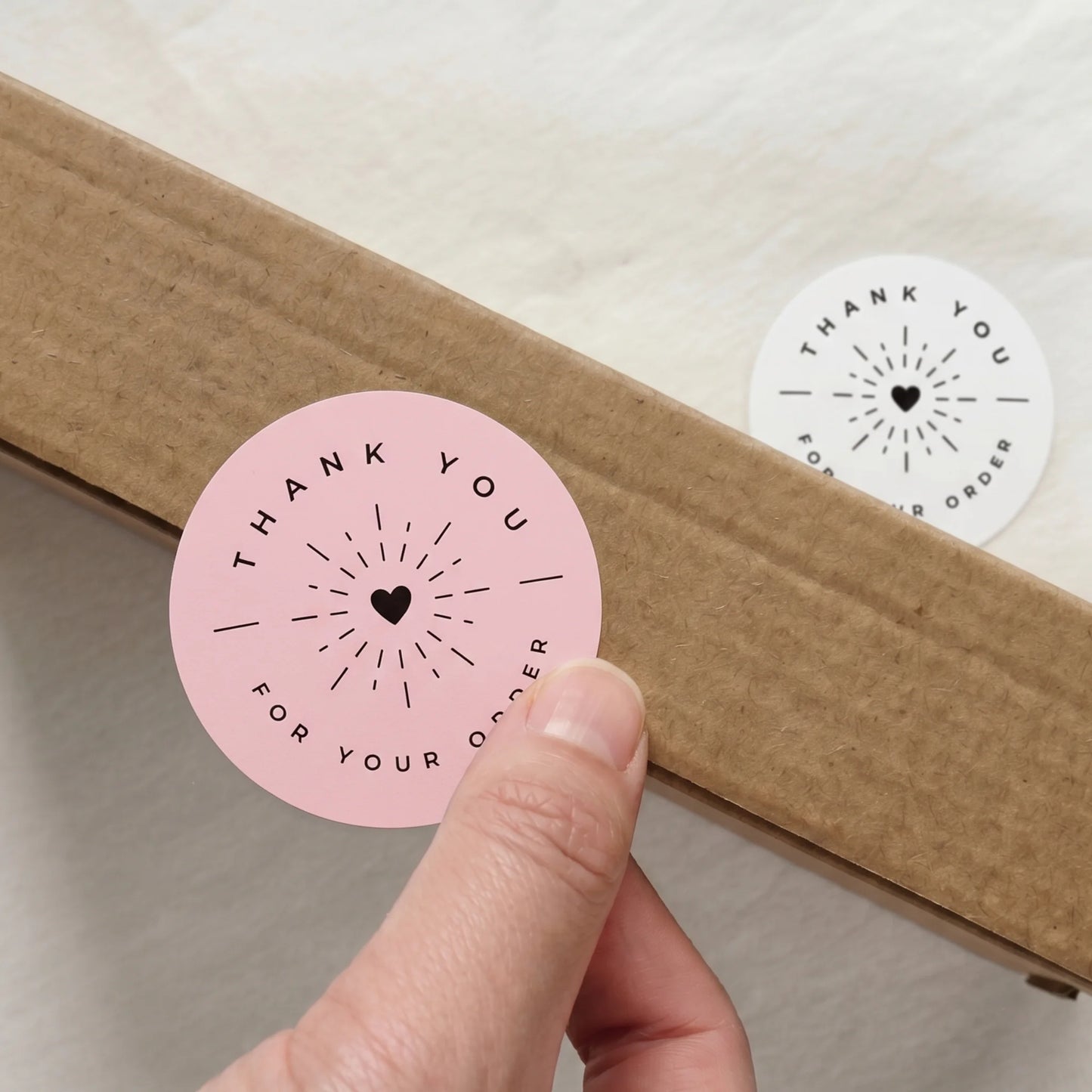 Round white sticker with 'Thank you for your order' text held over a cardboard box.