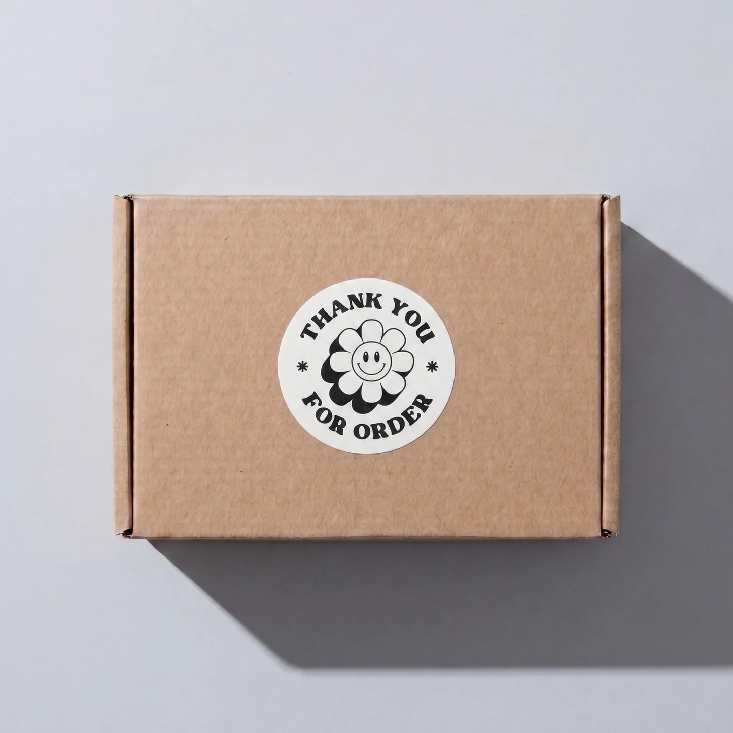 White 2-inch round Thank You sticker with black retro groovy daisy design, applied on a brown shipping box for small business order packaging by Varikit