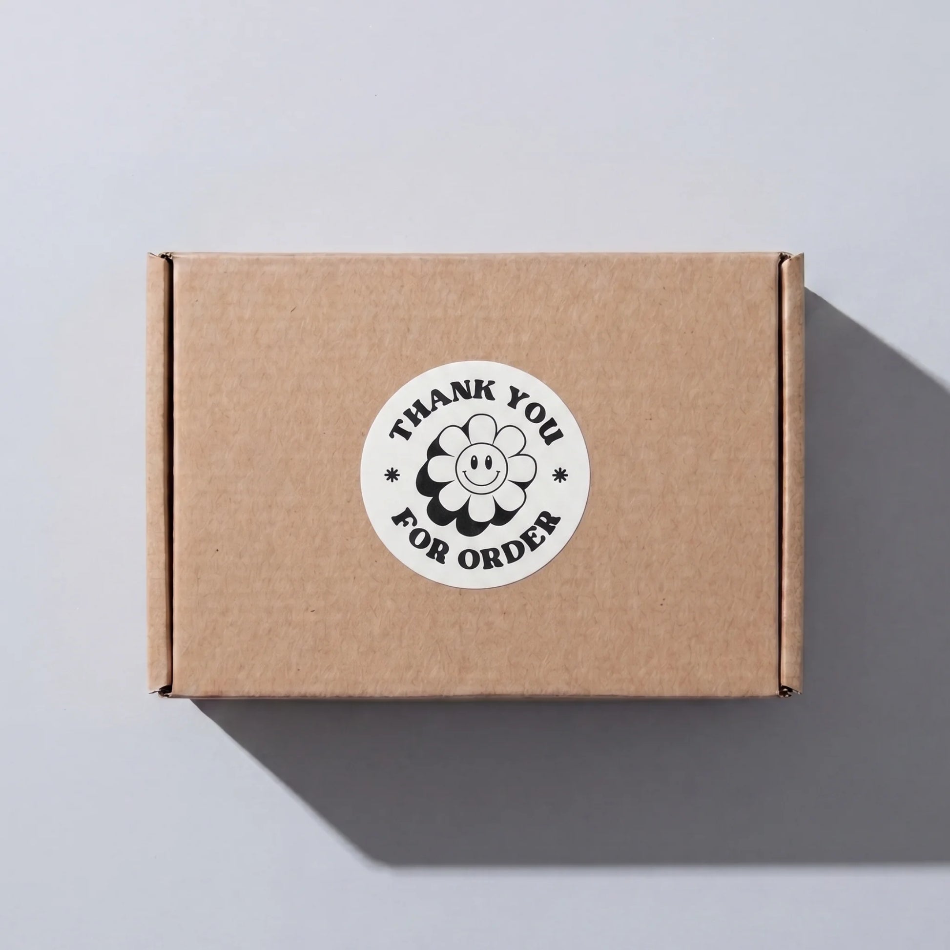 White 2-inch round Thank You sticker with black retro groovy daisy design, applied on a brown shipping box for small business order packaging by Varikit