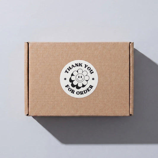 White 2-inch round Thank You sticker with black retro groovy daisy design, applied on a brown shipping box for small business order packaging by Varikit