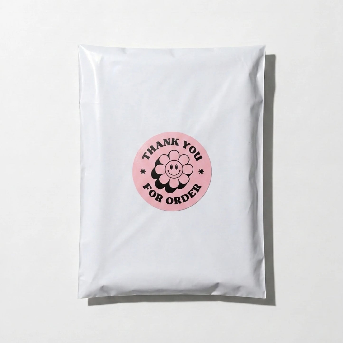 Pink 2-inch round Thank You sticker with black retro groovy daisy design, applied on a white poly mailer bag for small business packaging by Varikit