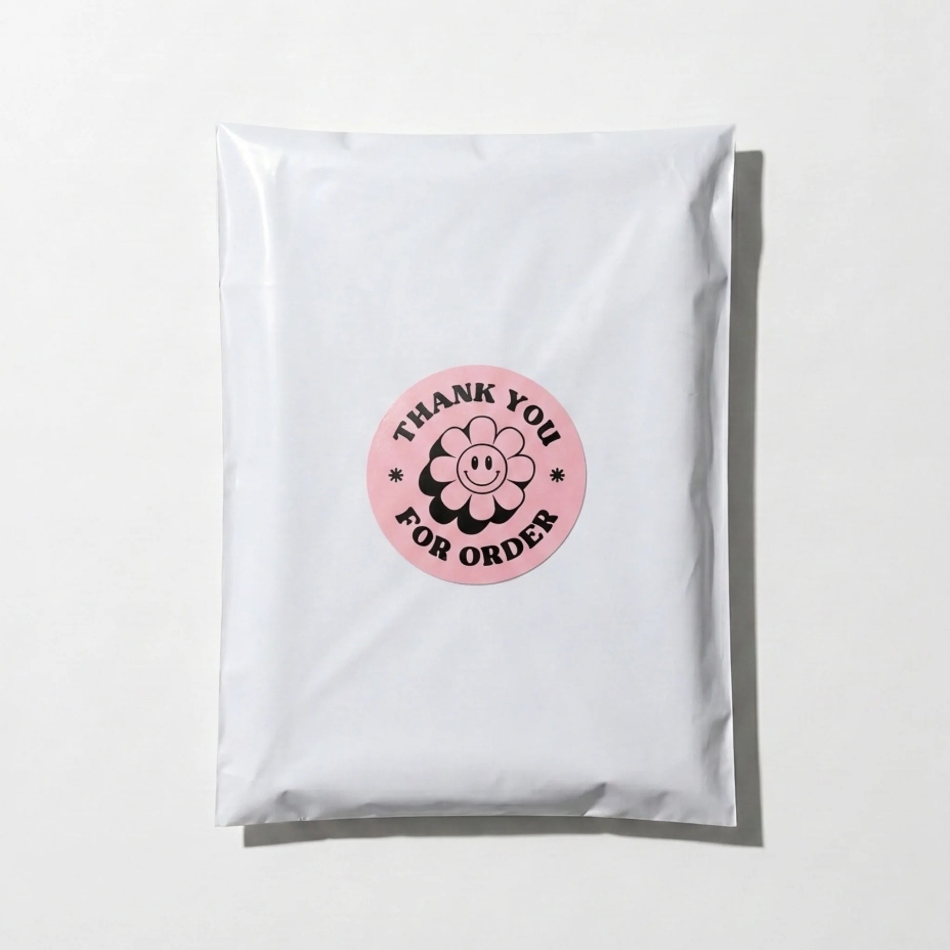 Pink 2-inch round Thank You sticker with black retro groovy daisy design, applied on a white poly mailer bag for small business packaging by Varikit
