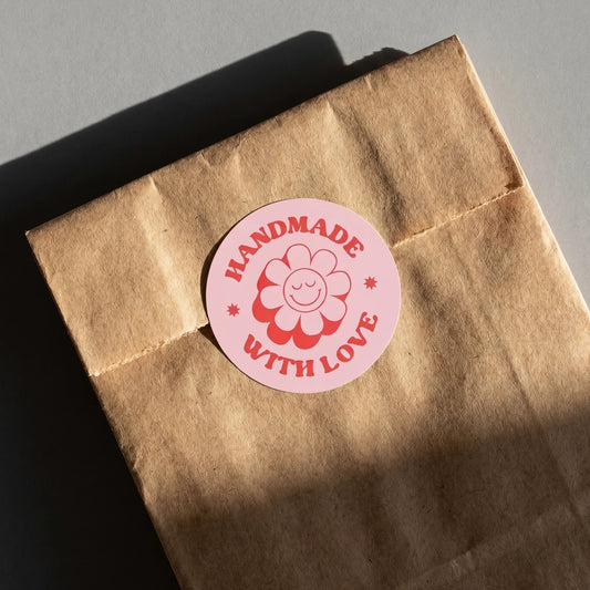 Hand holding a small lavender bubble mailer sealed with a Handmade With Love Retro Groovy Daisy Varikit sticker reading 'Handmade With Love'.