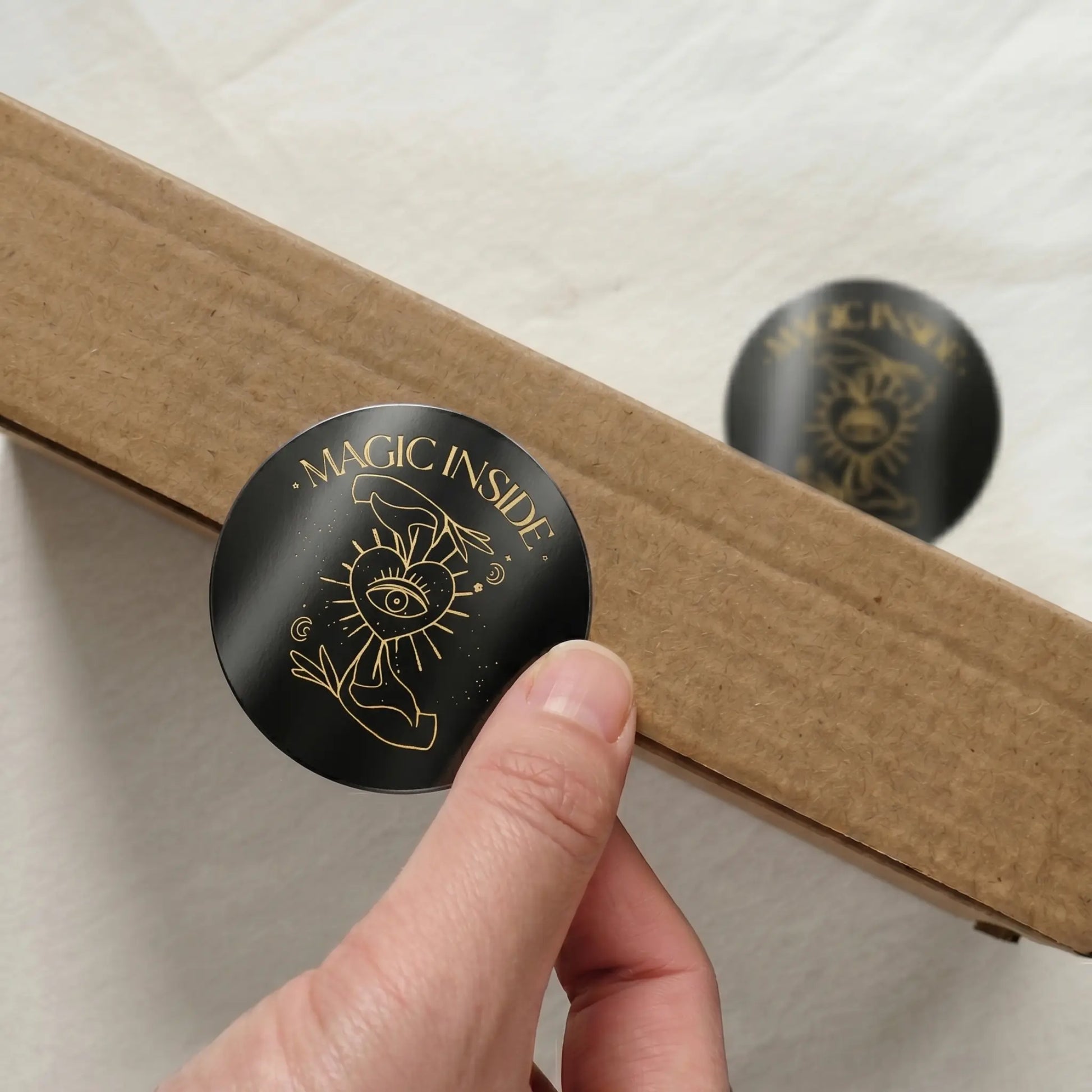 Black circular sticker with 'Magic Inside' text held by a hand against a cardboard box.