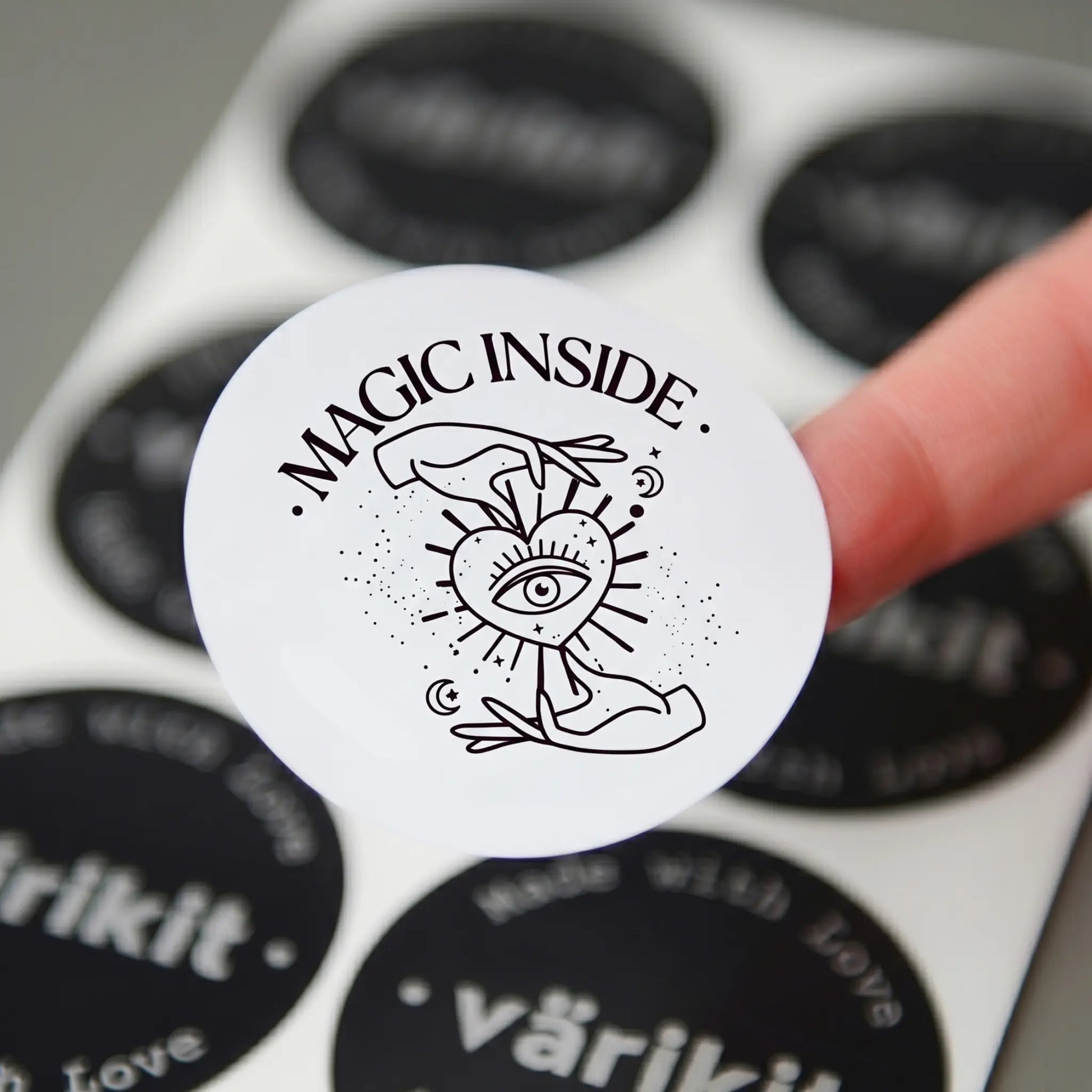 White sticker with 'Magic Inside' text and design held by a finger, with more stickers in the background.