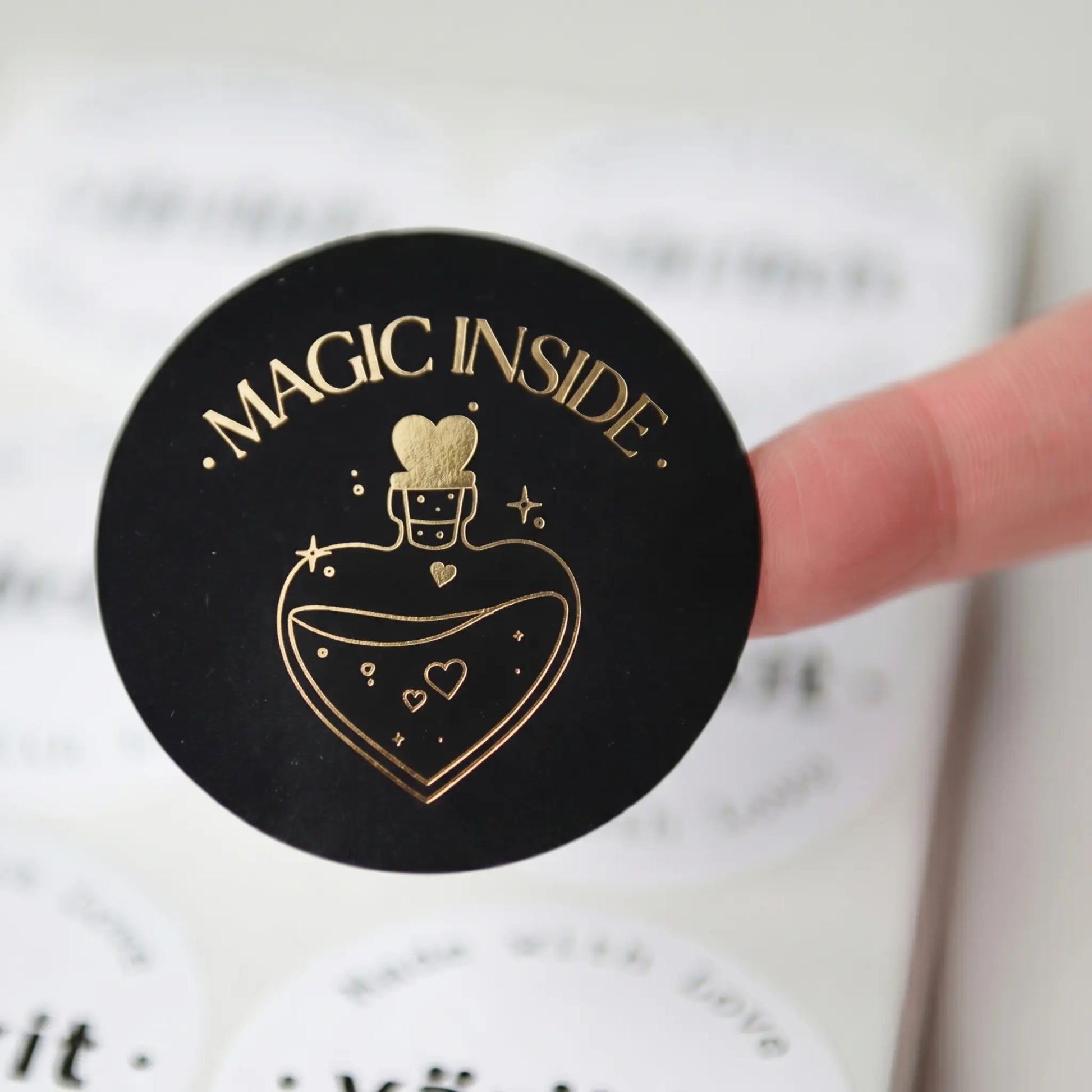 Black round container with gold foil text and design, held by a hand against a blurred background.