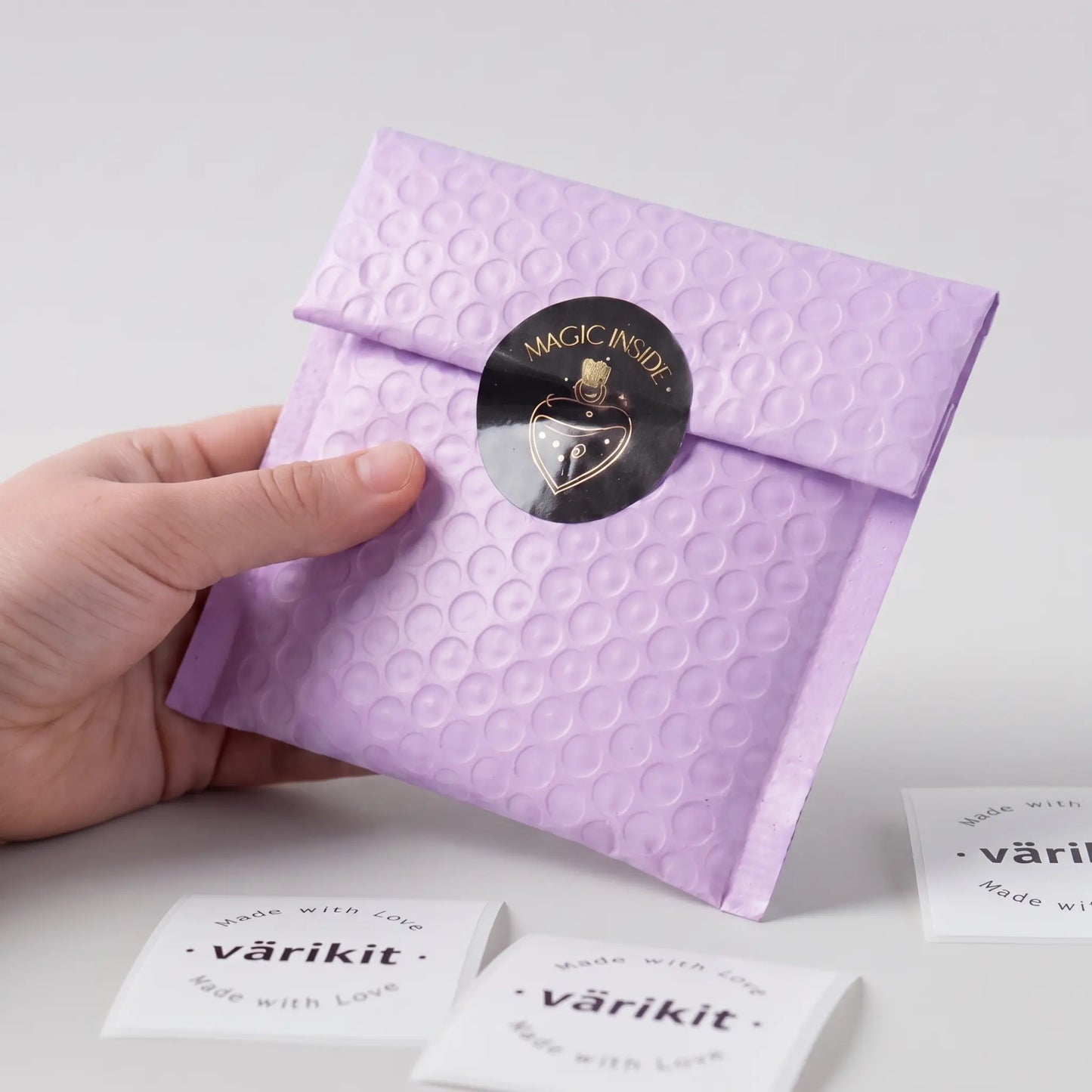 Hand holding a purple textured envelope with a black gold foil 'magic inside' sticker on a light gray background