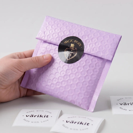 Hand holding a purple textured envelope with a black gold foil 'magic inside' sticker on a light gray background