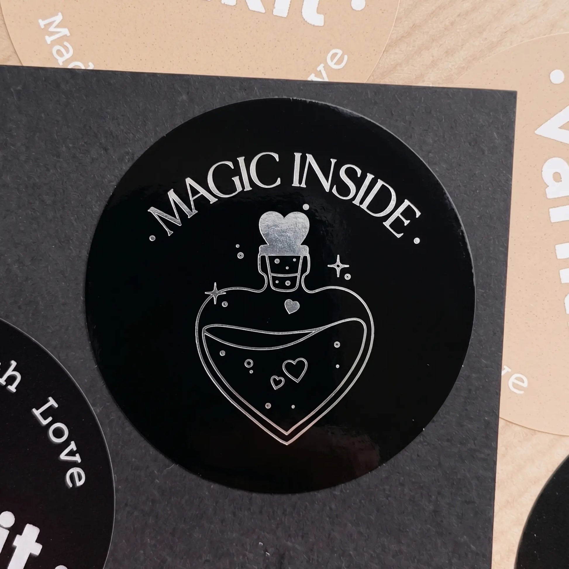 Black circular sticker with 'Magic Inside' text and potion design on a cardboard background.