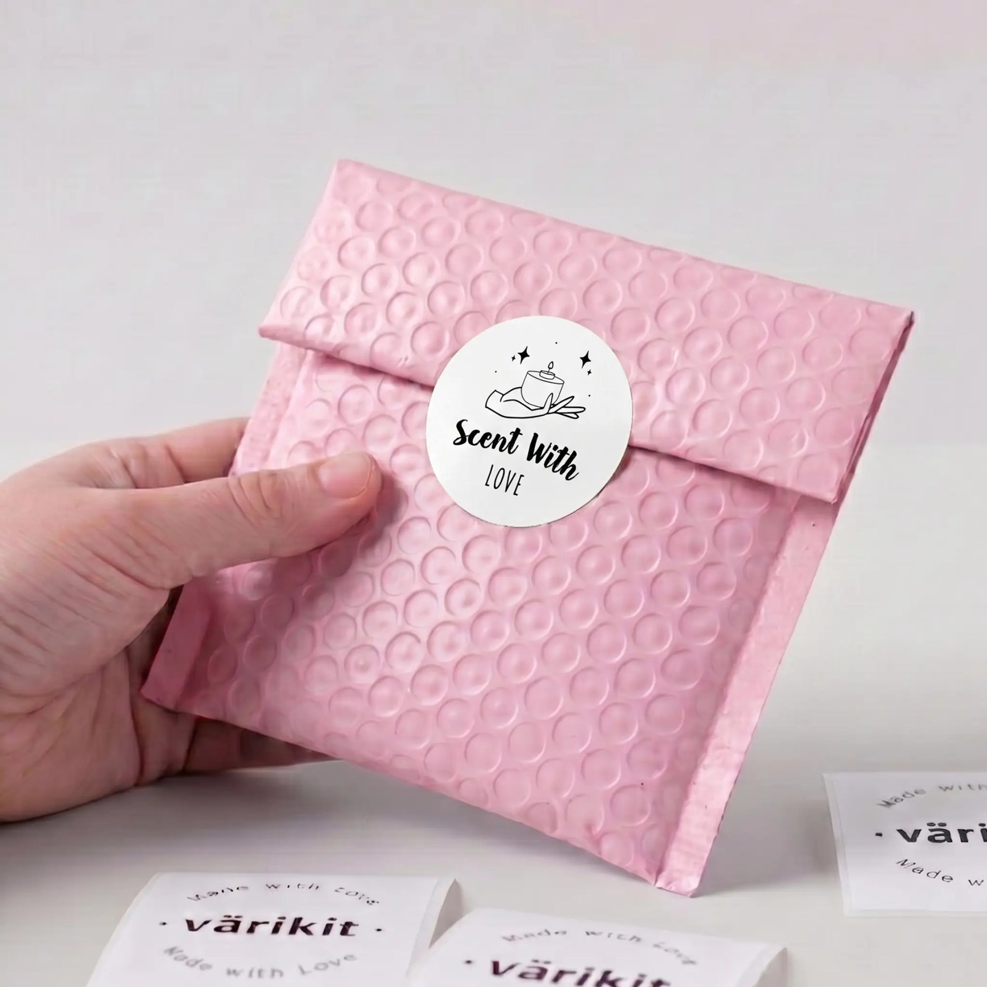 Hand holding a pink textured envelope with a black and white 'Scent With Love' label on a light gray background