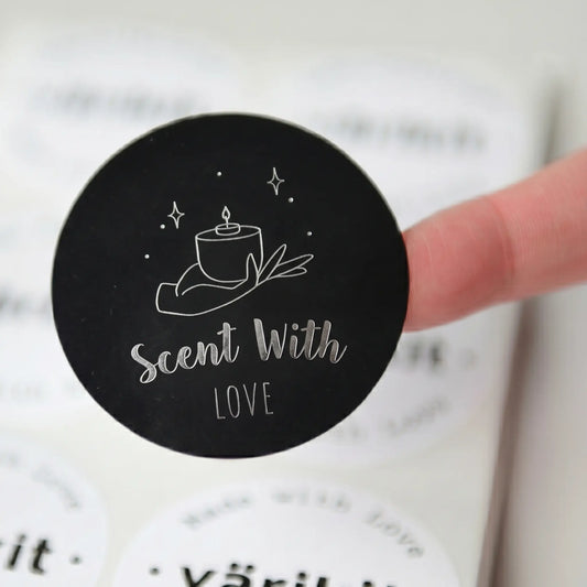 Round black sticker with 'Scent With Love' text and candle illustration held by a finger.