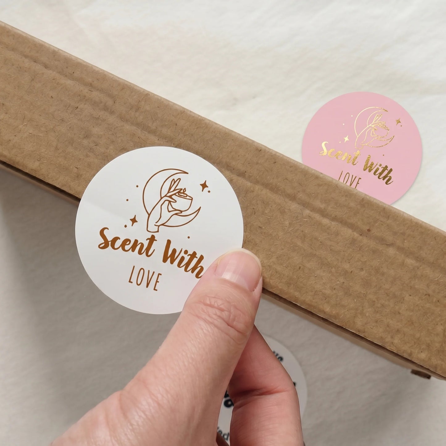 Hand placing a Scent With Love Mystic Candle Hand Varikit sticker reading 'Scent with Love' on a corrugated cardboard box.