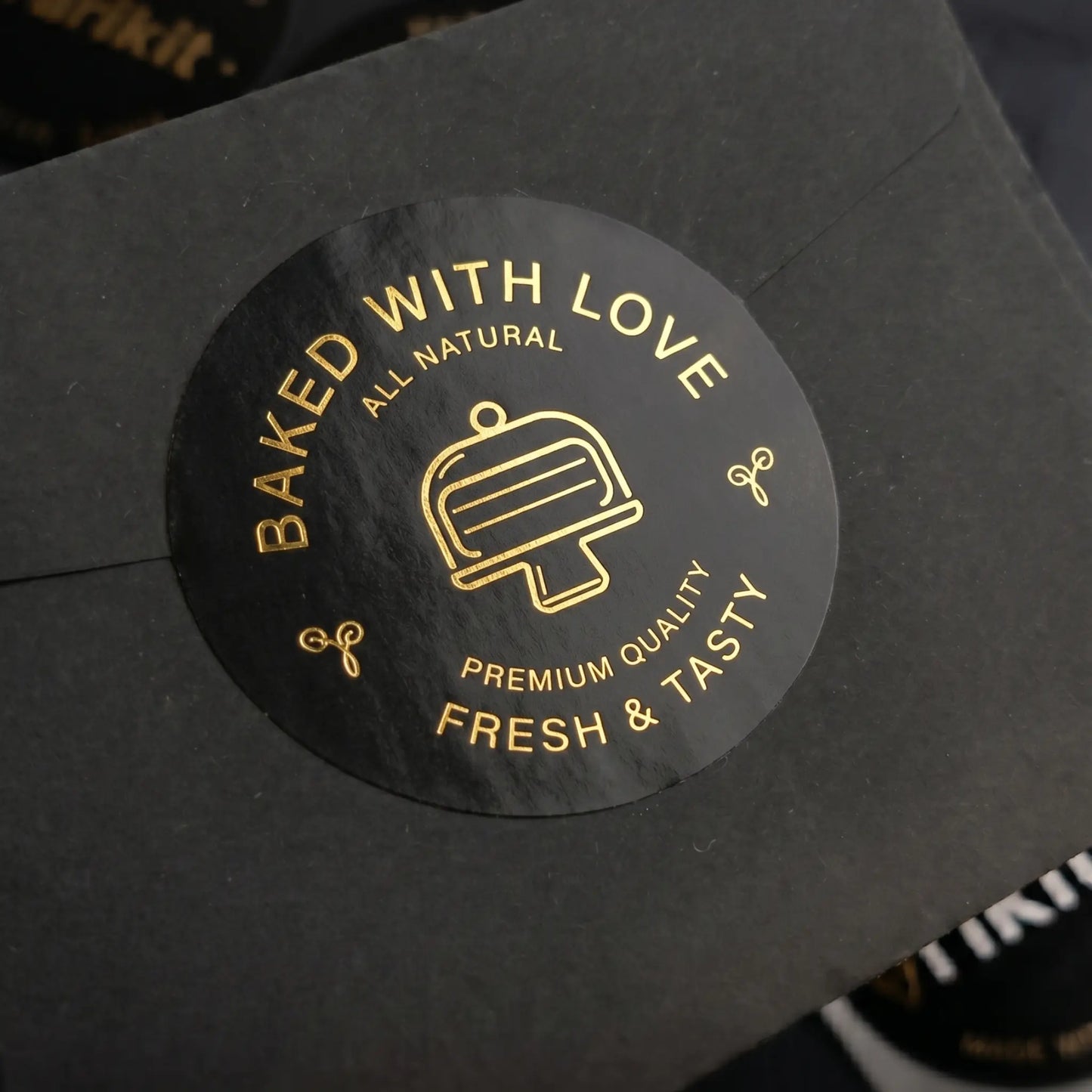 Black 'baked with love' label with gold foil text and cake stand graphic on a black background