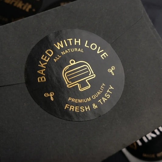 Black 'baked with love' label with gold foil text and cake stand graphic on a black background