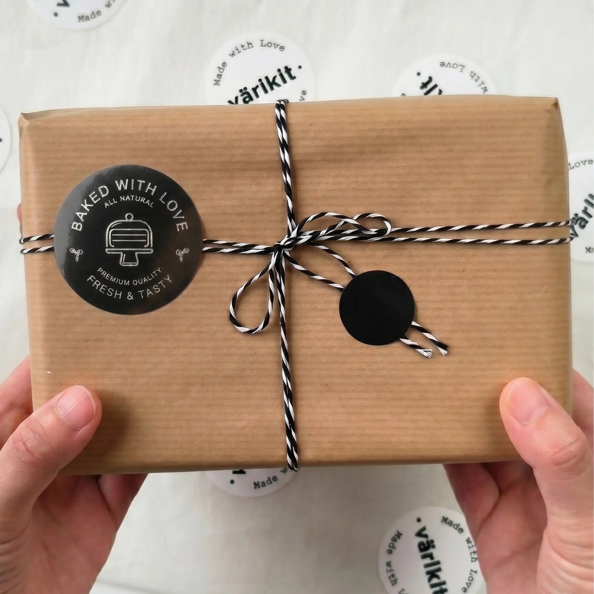 Brown paper-wrapped package with a black silver foil 'baked with love' label held by hands on a white background with 'varikit' branding.