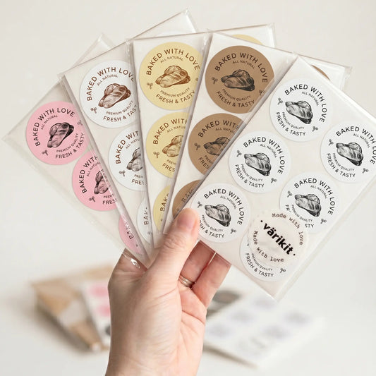 Hand holding a set of 'Baked with Love' stickers with various color schemes.