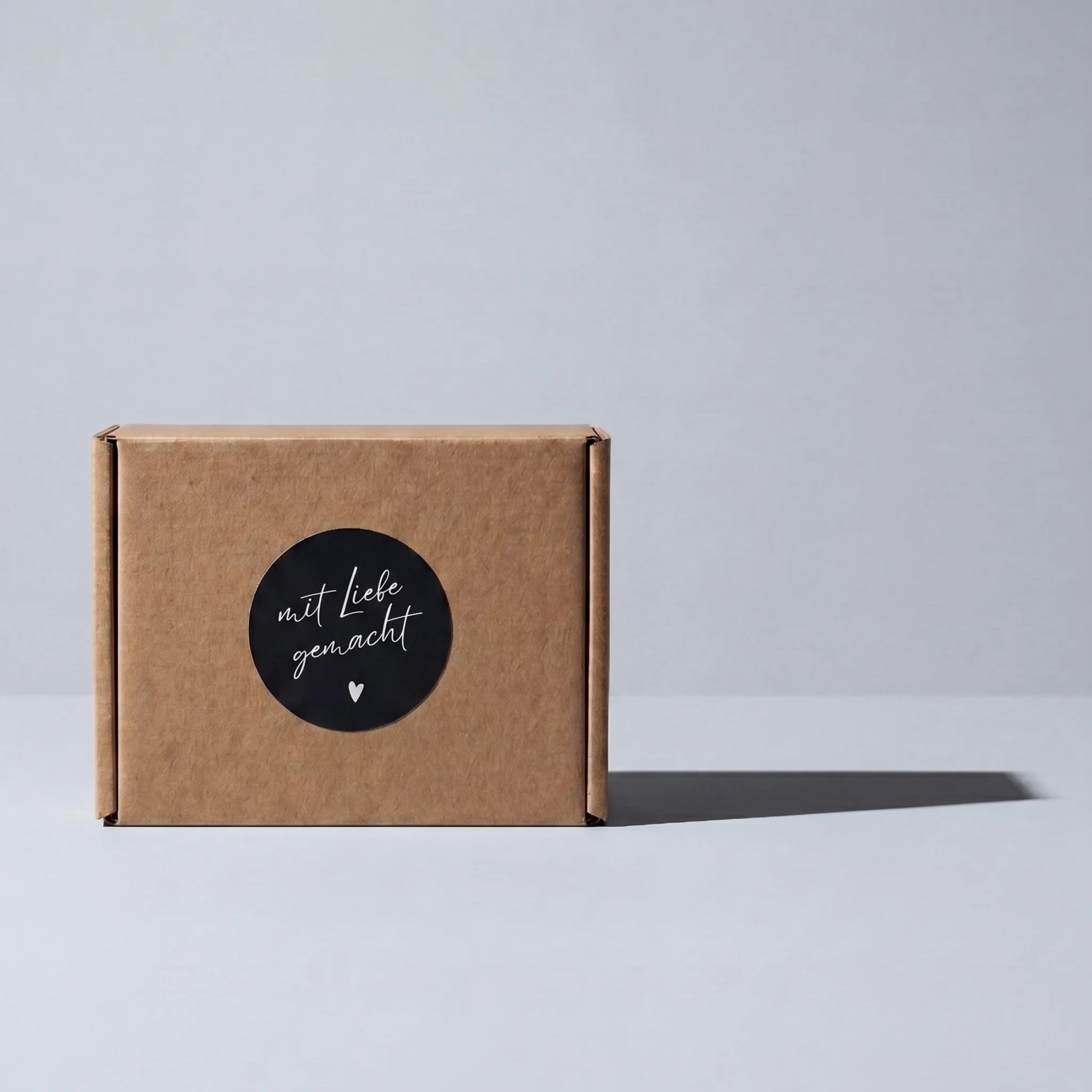 Brown cardboard box with a black 'mit Liebe gemacht' sticker on a light gray background