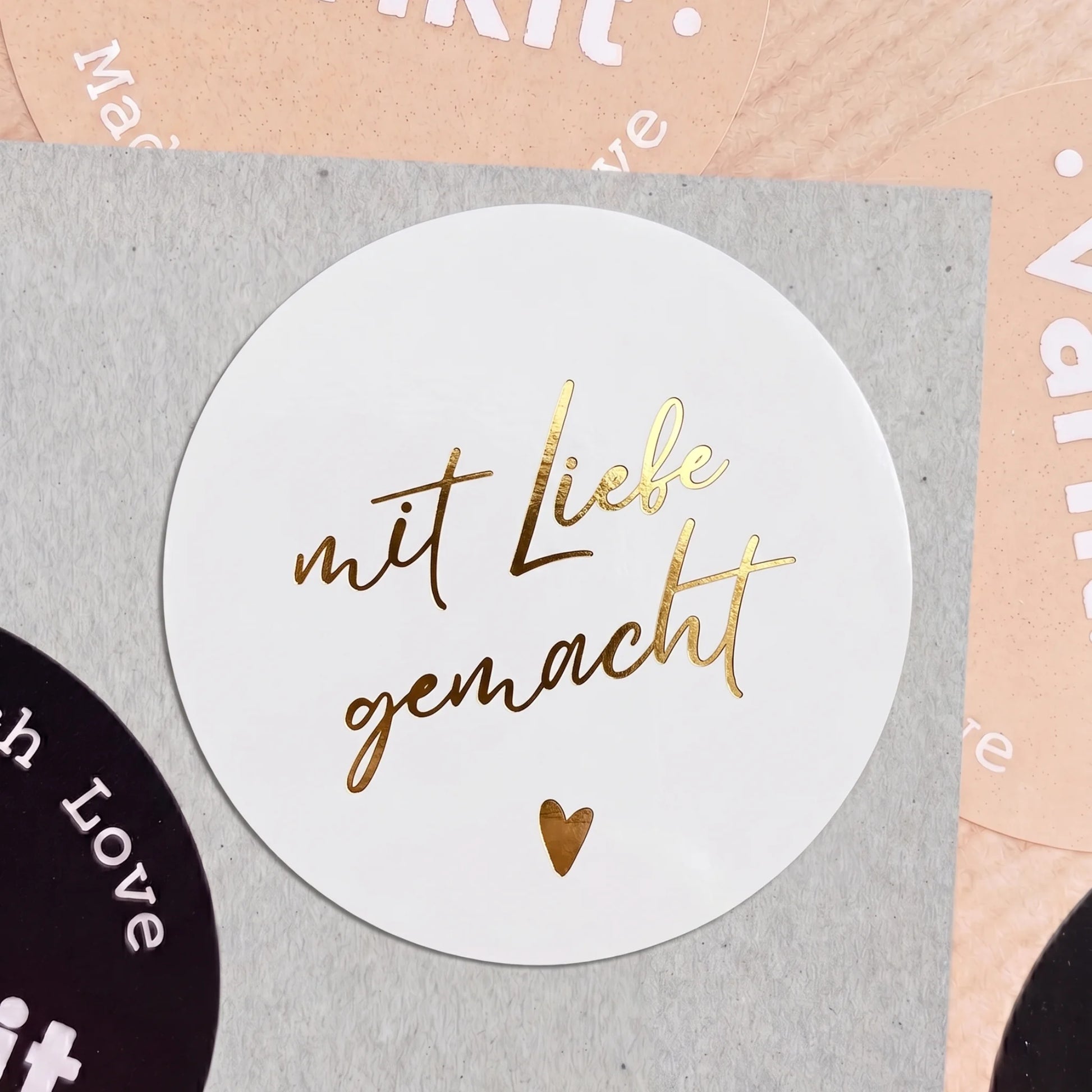 Round white sticker with gold text 'mit Liebe gemacht' on a gray background