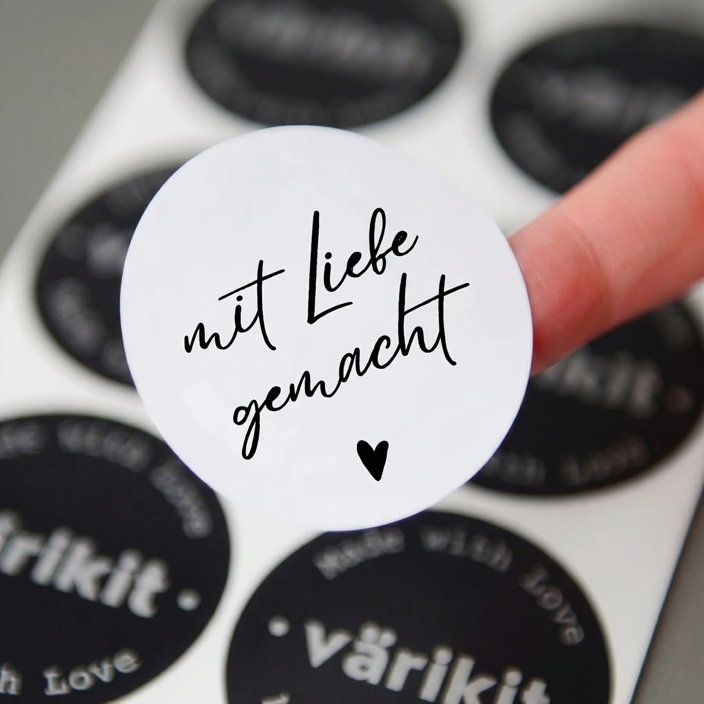 White sticker with 'mit Liebe gemacht' text held by a finger over black stickers with brand names.